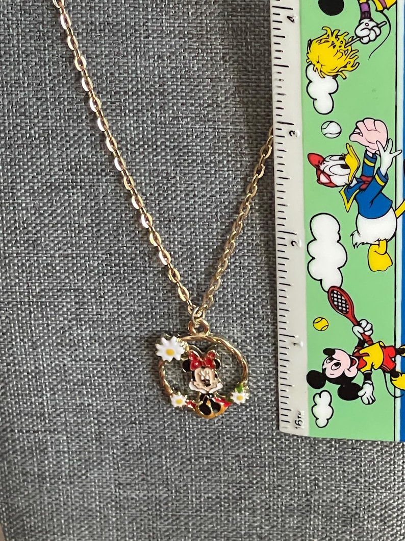 May include: Gold-colored chain necklace with a pendant featuring a cartoon character. The pendant is a circular design with a cartoon character in the center, surrounded by white flowers. The necklace is displayed against a gray background.