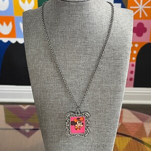 May include: A silver chain necklace with a silver frame pendant featuring a cartoon image of Minnie Mouse with a pink bow. The pendant is on a grey mannequin.