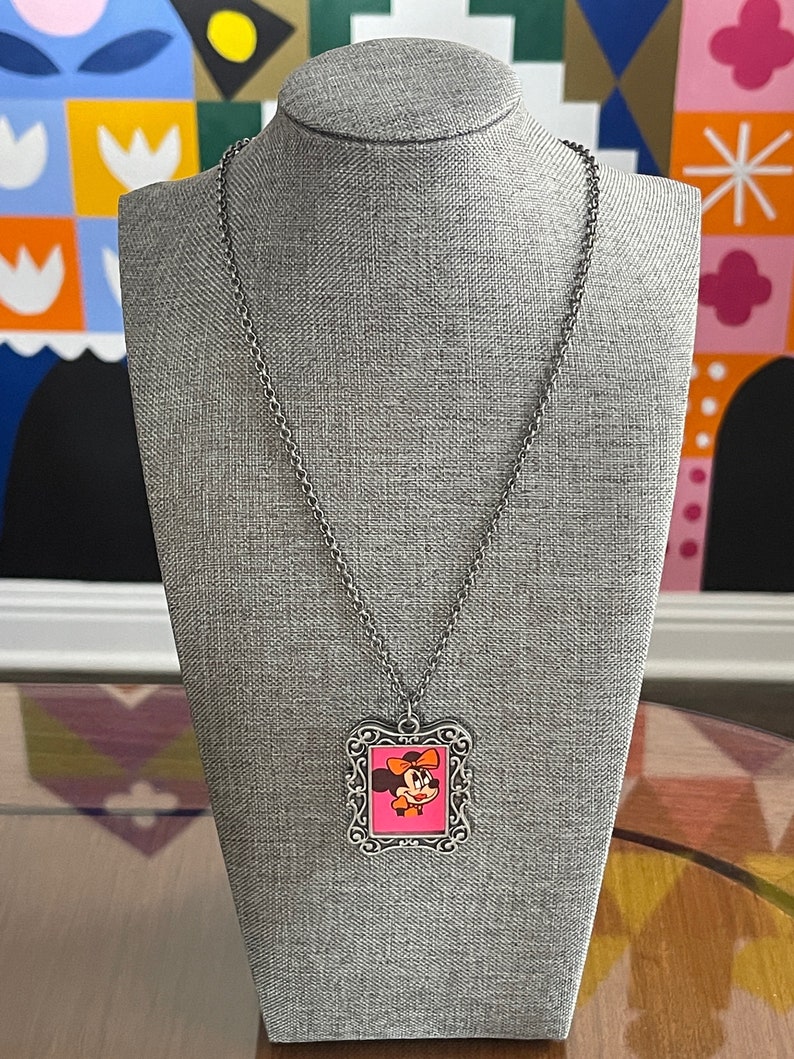 May include: A silver chain necklace with a silver frame pendant featuring a cartoon image of Minnie Mouse with a pink bow. The pendant is on a grey mannequin.