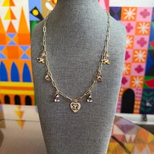 May include: Gold-toned chain necklace with Mickey Mouse charms and a heart pendant. The charms feature Mickey Mouse in various poses, with red, black, and white details. The heart pendant has a Mickey Mouse design. The necklace is displayed on a gray mannequin.