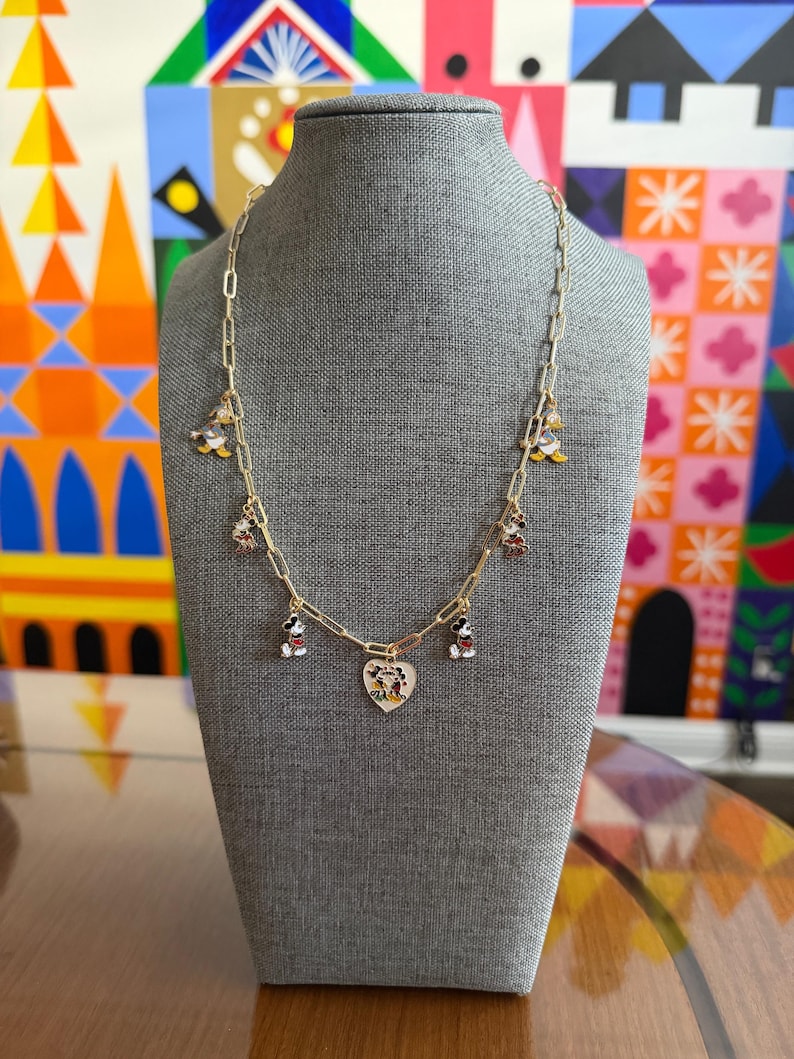 May include: Gold-toned chain necklace with Mickey Mouse charms and a heart pendant. The charms feature Mickey Mouse in various poses, with red, black, and white details. The heart pendant has a Mickey Mouse design. The necklace is displayed on a gray mannequin.