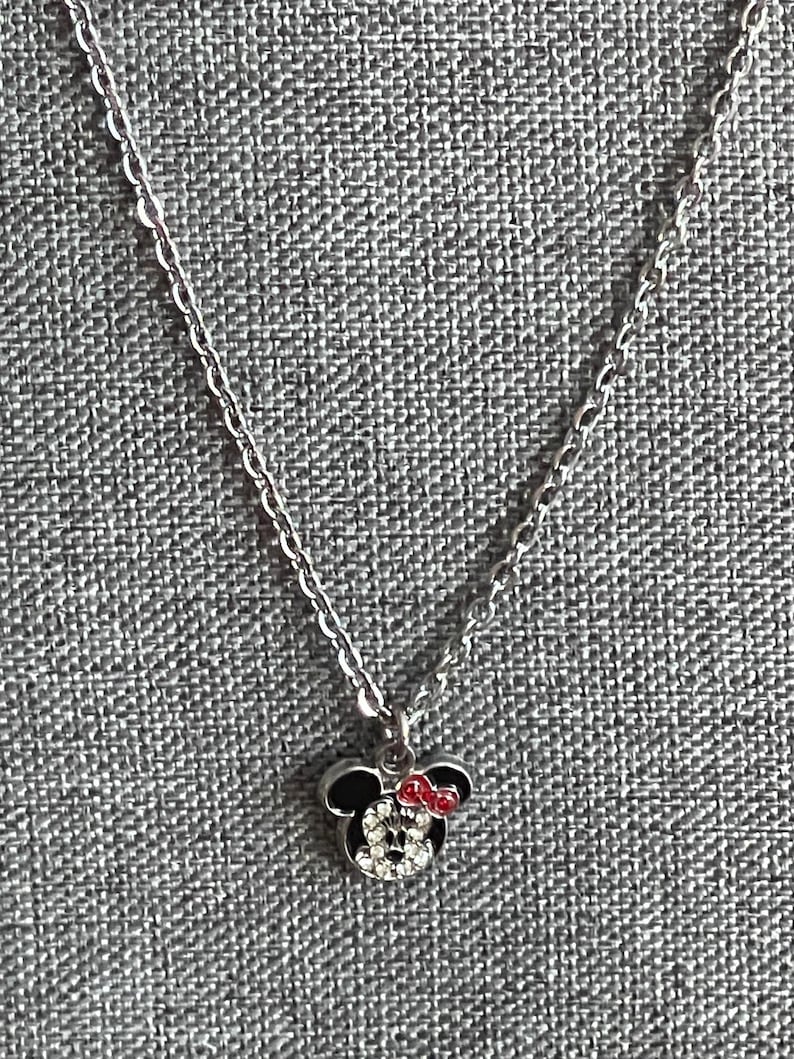 Pode incluir: Um colar prateado com um pingente da Minnie Mouse. O pingente apresenta uma cabe&ccedil;a preta com um la&ccedil;o vermelho e um rosto feito de cristais transparentes. O colar &eacute; exibido em uma superf&iacute;cie cinza texturizada.