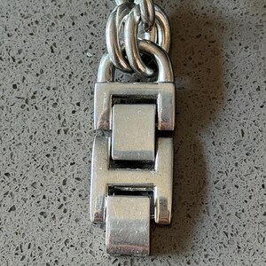 Puede incluir: Primer plano de un cierre y cadena de reloj plateados. El cierre es rectangular con un dise&ntilde;o articulado, y los eslabones de la cadena son ovalados. El metal tiene un acabado pulido, que refleja la luz.
