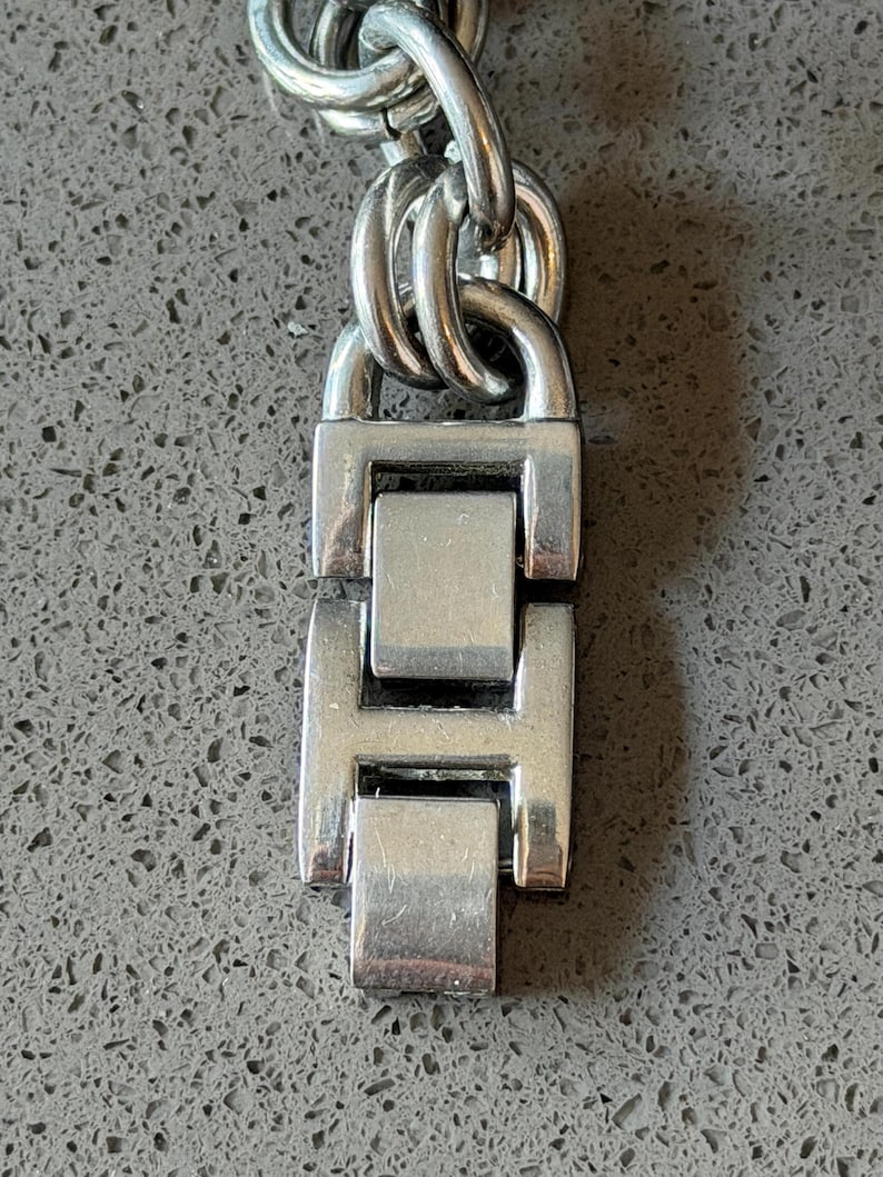 Puede incluir: Primer plano de un cierre y cadena de reloj plateados. El cierre es rectangular con un dise&ntilde;o articulado, y los eslabones de la cadena son ovalados. El metal tiene un acabado pulido, que refleja la luz.