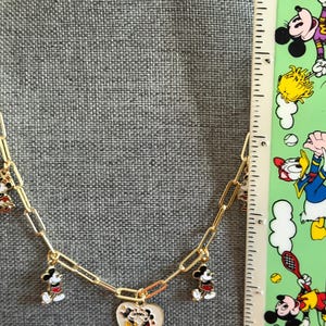 May include: Gold-toned chain necklace featuring Mickey Mouse charms. The necklace has a heart-shaped pendant with a Mickey and Minnie Mouse design. Additional charms include Mickey Mouse figures in red, black, and white. The necklace is displayed against a gray background.