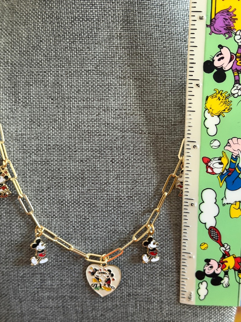 May include: Gold-toned chain necklace featuring Mickey Mouse charms. The necklace has a heart-shaped pendant with a Mickey and Minnie Mouse design. Additional charms include Mickey Mouse figures in red, black, and white. The necklace is displayed against a gray background.