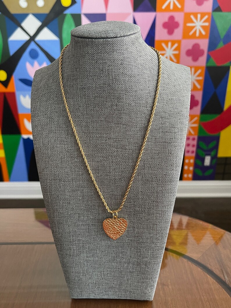May include: A gold chain necklace with a textured heart pendant. The necklace is displayed on a gray mannequin.