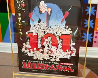 May include: A framed poster for the Walt Disney Classic 101 Dalmatians featuring Cruella de Vil in a blue coat above a large group of spotted puppies. The poster includes the text 101 Dalmatians, 99 2つの愛が、の助け合うやさしい心を育てた, and 101匹わんちゃん.