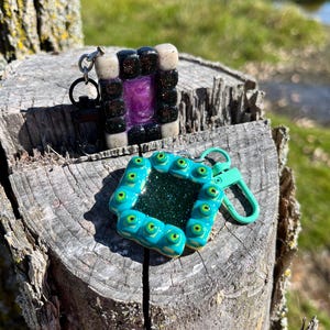 Könnte beinhalten: Zwei handgefertigte Schlüsselanhänger mit einem schwarz-weißen Quadrat mit einem violetten Zentrum und einem türkisfarbenen Quadrat mit grünen Augen um den Rand.