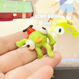 Puede incluir: Una figura verde con grandes ojos blancos y un pequeño corazón naranja en el pecho. La figura tiene un sombrero rojo con una hélice negra. El texto "BING BONG" y "pick up" son visibles en el fondo.