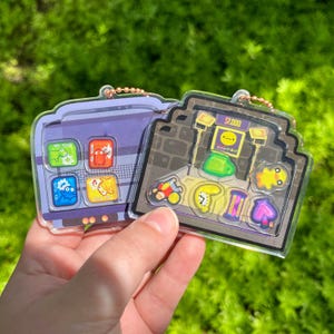 Puede incluir: Dos llaveros de acrílico transparente con enganches de cadena dorados. Un llavero es morado con iconos de aplicaciones coloridos. El otro llavero es un diseño de juego de arcade marrón y gris con varios iconos relacionados con juegos.