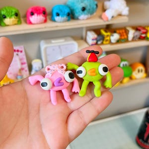 Puede incluir: Dos figuras de arcilla hechas a mano, una rosa con un lazo y otra verde con un sombrero de hélice, se sostienen en una mano. Las figuras tienen ojos grandes y saltones. Otras pequeñas criaturas hechas a mano son visibles en el fondo.