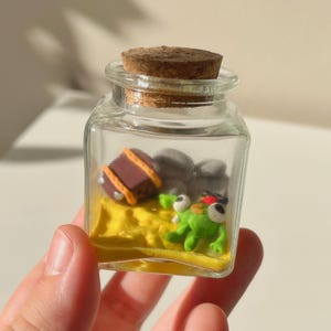 Puede incluir: Un pequeño frasco de vidrio transparente con un tapón de corcho, que contiene una escena en miniatura. En el interior, hay una base amarilla, un cofre del tesoro marrón, rocas grises y una figura de rana verde con un sombrero rojo.
