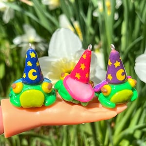 Può includere: Tre figurine di rana verde che indossano cappelli da mago colorati con stelle e una mezzaluna. I cappelli sono blu, rosa e viola.