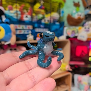 May include: A small, handcrafted figurine of a blue octopus with white speckles and a smooth, glossy finish. The creature features large, dark, round eyes and a white underbelly, with several curved tentacles extending outward from its body.