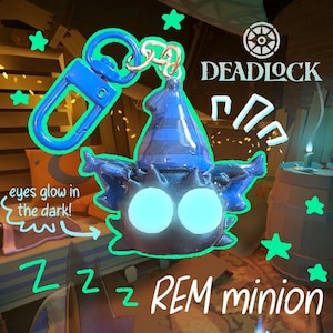 Peut inclure: Un porte-clés REM minion bleu et noir avec des yeux brillants. Le porte-clés a un clip bleu et un anneau doré. Le minion a un chapeau bleu et est entouré d'étoiles vertes. Le texte "DEADLOCK" est visible.
