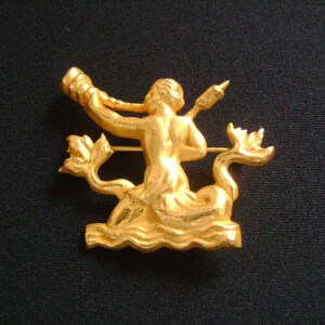 May include: A gold-toned brooch featuring a mermaid figure playing a horn, with a stylized wave design at the base.