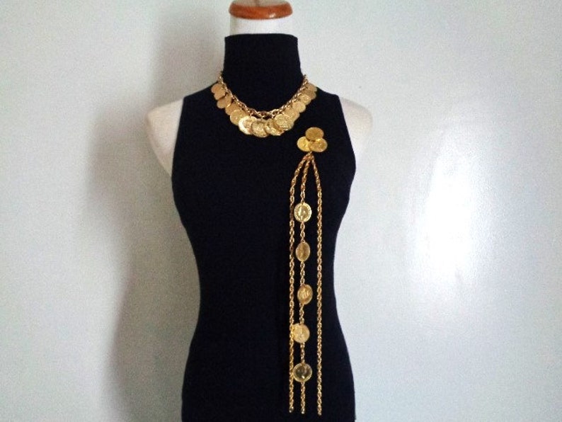 May include: A gold-tone necklace and matching brooch with a chain and coin design. The necklace is layered and the brooch hangs from a long chain.