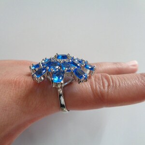 May include: A silver ring with a cluster of blue oval-shaped gemstones. The ring is set on a finger.