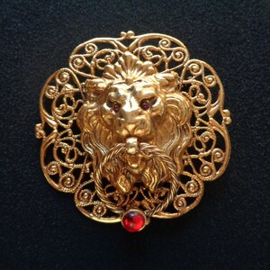 May include: Gold-toned lion head brooch with a red jewel in the centre, surrounded by a filigree design.