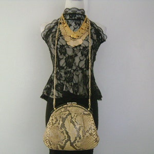 May include: A snakeskin clutch purse with a gold clasp and chain strap. The purse is on a mannequin wearing a black lace top and a gold necklace.