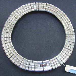Puede incluir: Un collar plateado hecho de cuadrados de metal unidos. El collar es de estilo gargantilla y tiene un cierre de broche.