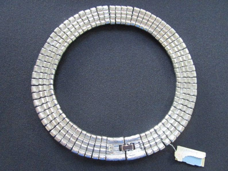 Puede incluir: Un collar plateado hecho de cuadrados de metal unidos. El collar es de estilo gargantilla y tiene un cierre de broche.