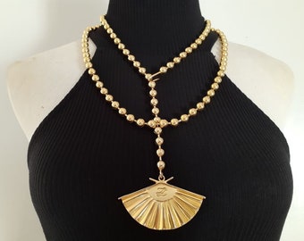 May include: A gold-toned necklace with a large, fan-shaped pendant. The necklace is made of round beads and has a layered design.