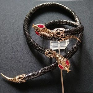 May include: A black and gold snake-shaped necklace with red detailing. The snake's head and tail feature gold metal and red gemstones. The necklace is displayed on a clear stand against a dark background.