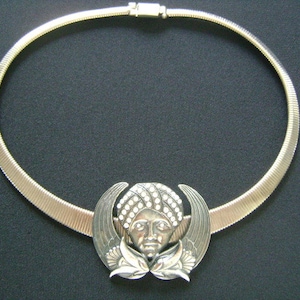 May include: A silver necklace with a pendant featuring a detailed face with a turban. The pendant is surrounded by wings.