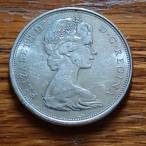 May include: A silver coin featuring a profile of a woman wearing a crown, with the inscription "ELIZABETH II D.G. REGINA" around the edge. The coin is set against a wooden surface.