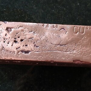 May include: A copper bar with a textured surface. The bar is stamped with "4.6 LB" and "CU 999". The copper has a reddish-brown color.
