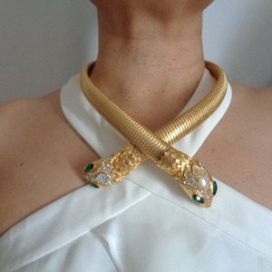 May include: A gold snake necklace with a textured, ribbed design. The snake heads are adorned with clear and iridescent crystals, and green and white gemstones. The necklace is worn over a white top.