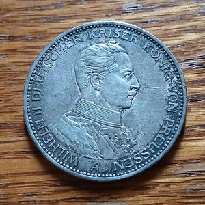 May include: A silver coin featuring a profile of a man with a mustache, surrounded by text. The coin is likely a historical artifact, possibly a commemorative piece or currency. The coin is on a wooden surface.