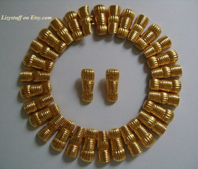 May include: A gold-tone necklace and matching earrings. The necklace is made of a series of linked, textured, cylindrical beads. The earrings are simple, with a single textured, cylindrical bead.