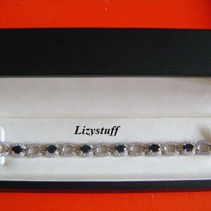 May include: A silver bracelet with oval-shaped blue gemstones and smaller clear gemstones. The bracelet is in a black and white box with the word "Lizystuff" printed on the inside.