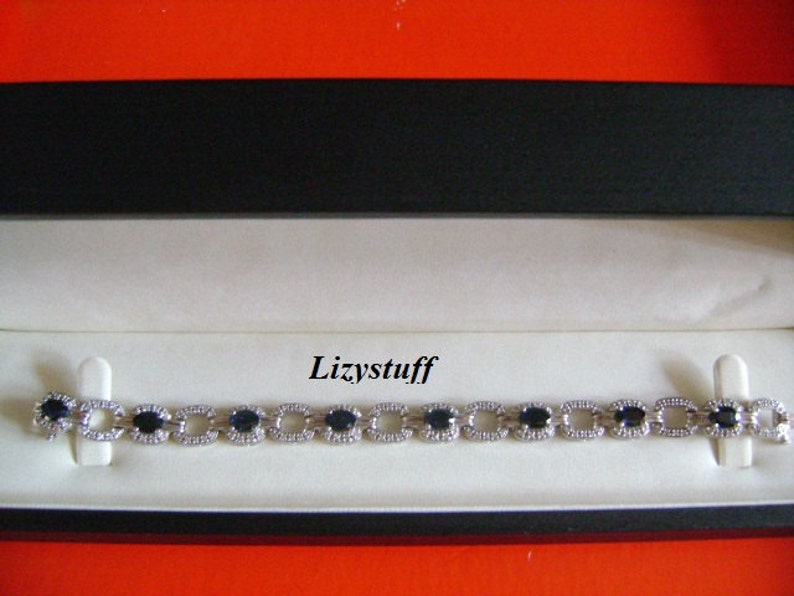 May include: A silver bracelet with oval-shaped blue gemstones and smaller clear gemstones. The bracelet is in a black and white box with the word "Lizystuff" printed on the inside.