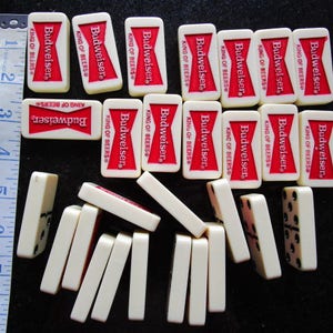 May include: A set of dominoes with a Budweiser King of Beers theme. The dominoes are white with black dots and feature a red and white Budweiser logo.