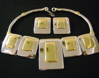 Puede incluir: Un collar de plata y oro con un diseño geométrico. El collar está formado por cinco piezas rectangulares, cada una con un centro dorado y un borde plateado. El collar está unido a una cadena de plata.