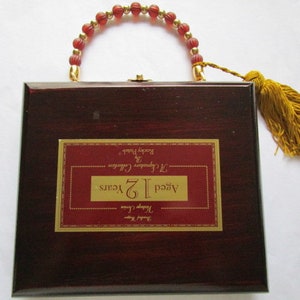 Op de afbeelding: Een houten doos met een rode afwerking en een gouden label met de tekst "Aged 12 Years,  J.C.  Cigars,  Collectors Edition,  Rocky Patel". De doos heeft een rode kralenhendel en een gouden kwast.