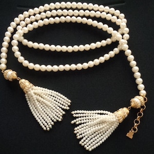 Puede incluir: Un collar largo de perlas blancas con borlas doradas en cada extremo. El collar está hecho de pequeñas perlas redondas y está enhebrado en una cadena dorada.