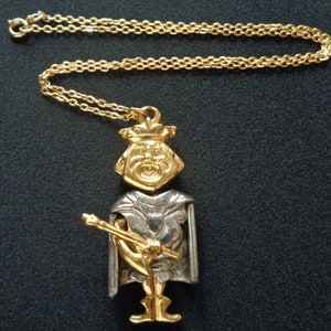 May include: A gold and silver pendant necklace featuring a stylized figure wearing a crown and holding a staff. The figure is wearing a long robe and has a detailed face.