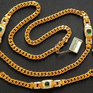 May include: A gold-toned jewelry set featuring a necklace, bracelet, and earrings. The necklace and bracelet have a curb chain design with emerald-colored center stones and clear accent stones. The earrings have a similar design. The set is displayed on a dark surface.
