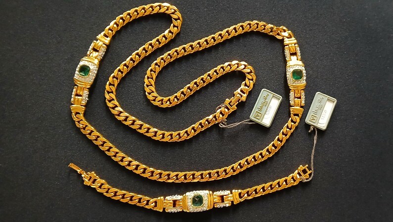 May include: A gold-toned jewelry set featuring a necklace, bracelet, and earrings. The necklace and bracelet have a curb chain design with emerald-colored center stones and clear accent stones. The earrings have a similar design. The set is displayed on a dark surface.