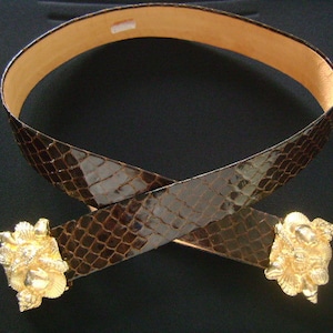 May include: A brown leather belt with a snakeskin pattern and two gold-toned buckles. The buckles are ornate and feature a design of seashells.