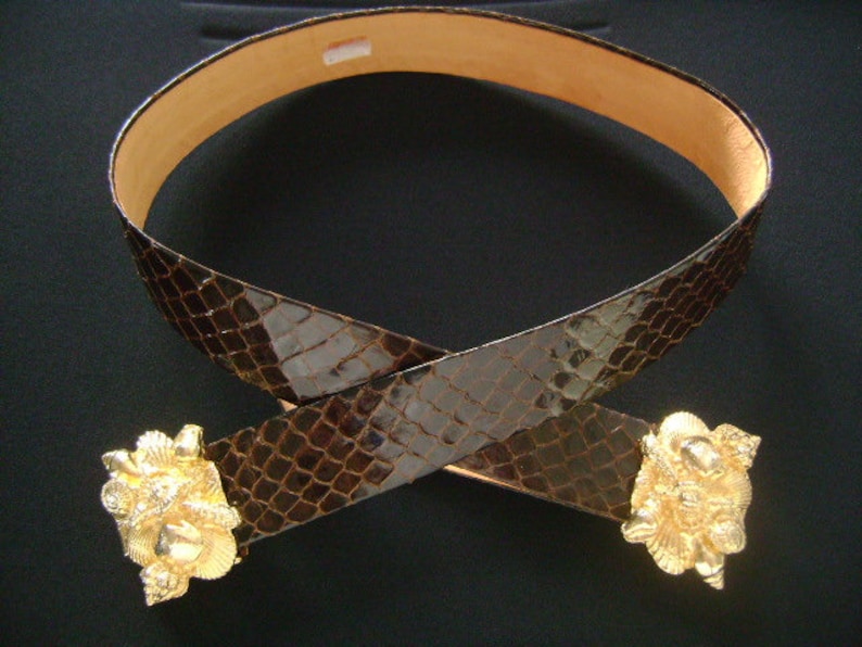May include: A brown leather belt with a snakeskin pattern and two gold-toned buckles. The buckles are ornate and feature a design of seashells.