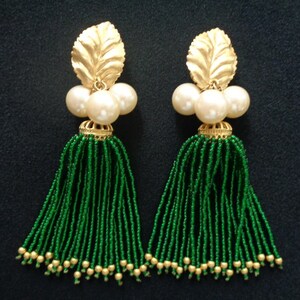 May include: A pair of dangling earrings featuring a gold leaf design at the top, followed by three white pearls, a gold filigree, and a long green beaded tassel. The tassels are accented with small gold beads at the bottom.