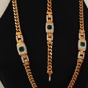 May include: Gold-toned jewelry set featuring a necklace and bracelet. The necklace and bracelet have a chain link design with emerald-green square center stones surrounded by clear crystals. The jewelry is displayed on a black background.