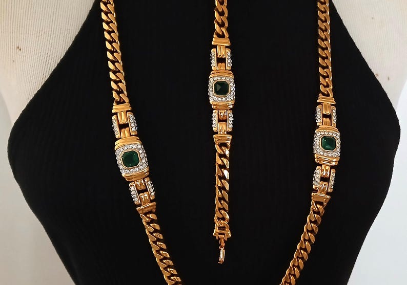 May include: Gold-toned jewelry set featuring a necklace and bracelet. The necklace and bracelet have a chain link design with emerald-green square center stones surrounded by clear crystals. The jewelry is displayed on a black background.