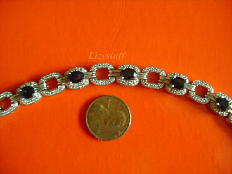 May include: A silver bracelet with square links and dark blue oval gemstones. The bracelet is shown next to a US penny for size comparison.